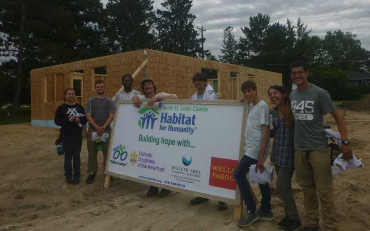 A group of young people pose for a photo around a habitat for humanity sign. Behind them is the frame of a house.